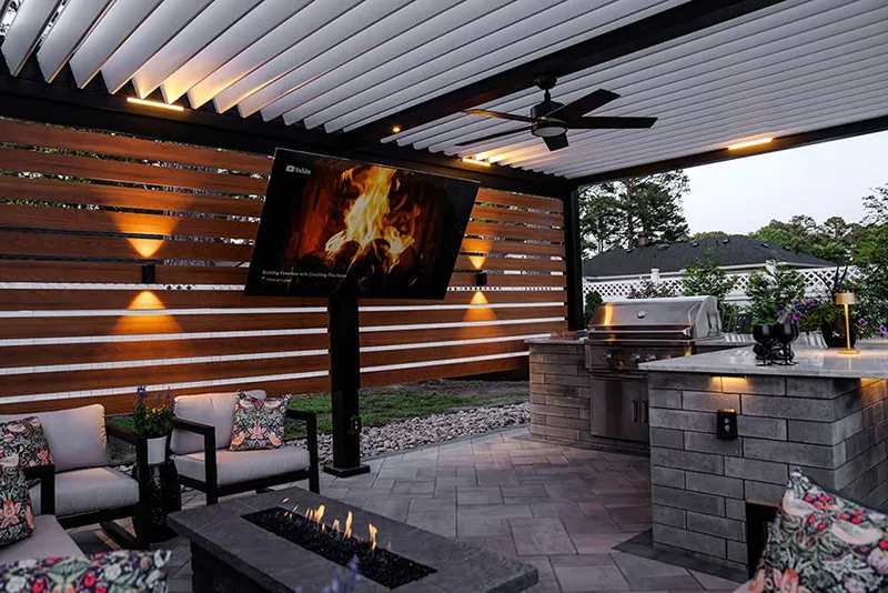 Custom-designed pergola with automated louvered roof in aluminum: Model R-BLADE with additional privacy wall where stands the outdoor TV, fan and LED lights - Made in the USA by Azenco Outdoor.