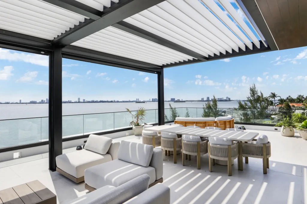 Waterfront dining and lounging area shaded by a black Azenco R-BLADE™ pergola with white louvered roof and glass railing.