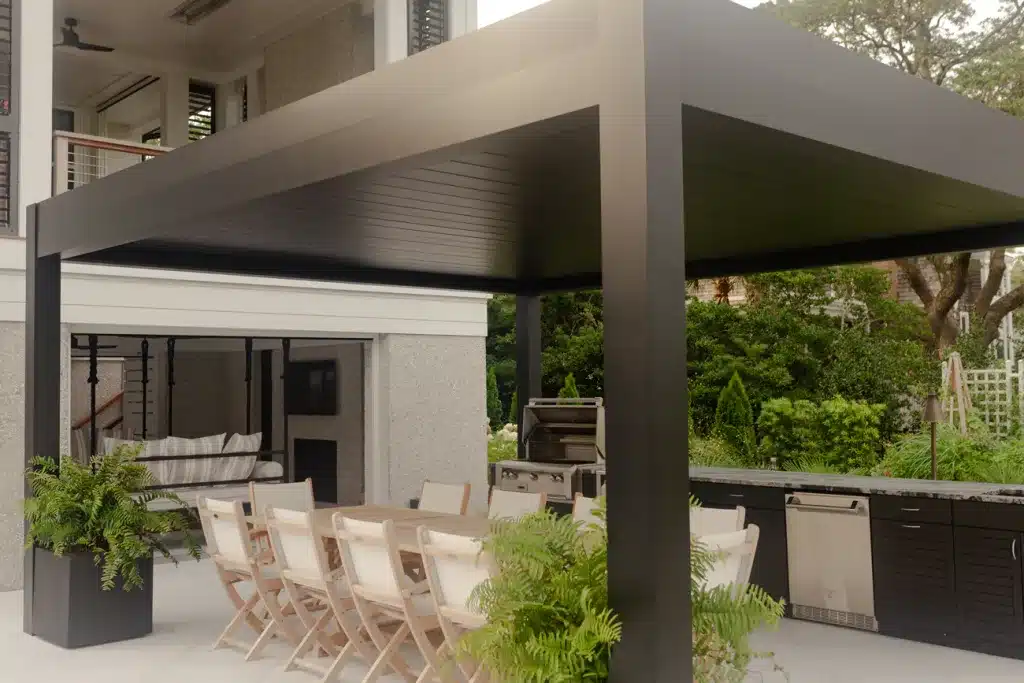 Modern black R-blade pergola providing shade over a light wood outdoor dining table and an integrated outdoor kitchen with dark cabinetry.