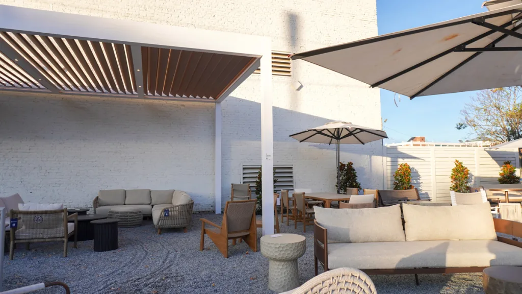 A wide view of the Azenco outdoor living space at Broad Hall, High Point. A white-framed pergola with wood-look R-Blade louvered roofing covers a seating area with a light beige sectional sofa and woven furniture on a gravel patio, next to a large white market umbrella.
