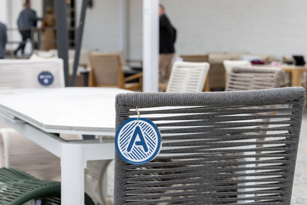 A close-up, shallow-depth-of-field shot of high-quality Azenco outdoor furniture at Broad Hall High Point. The back of a grey woven rope chair has a blue Azenco logo tag hanging from it, set next to a white slatted dining table. People are visible in the blurred background.