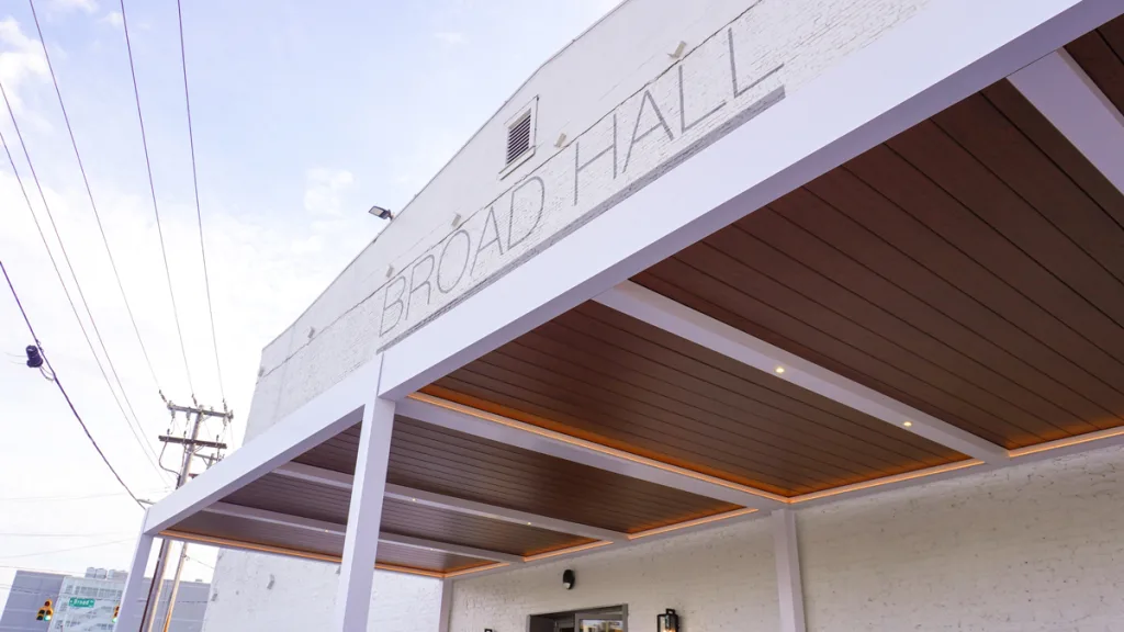 A low-angle exterior view showing the classic white brick facade of "BROAD HALL" and the white Azenco R-Blade motorized louvered pergola entrance. The wood-look ceiling panels are equipped with subtle, warm LED lighting, highlighting the installation at High Point, NC.