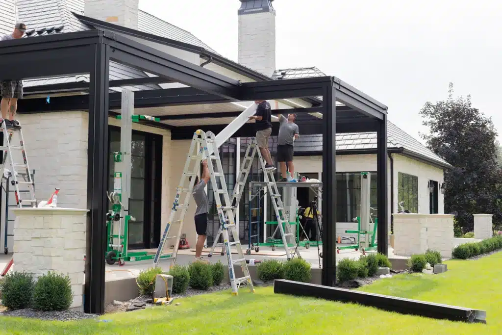 Installation of a black Azenco R-BLADE™ louvered pergola by a crew on ladders, attaching the roof panels to the aluminum frame.