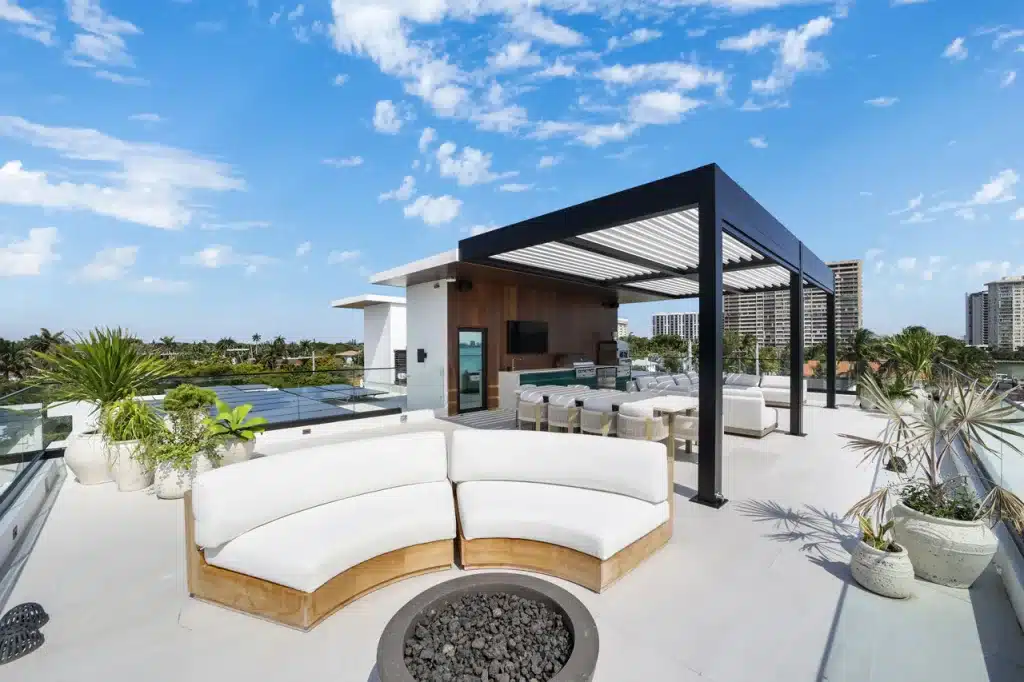 Rooftop lounge featuring a black Azenco R-BLADE™ pergola, curved sectional seating around a fire pit, and outdoor kitchen area.