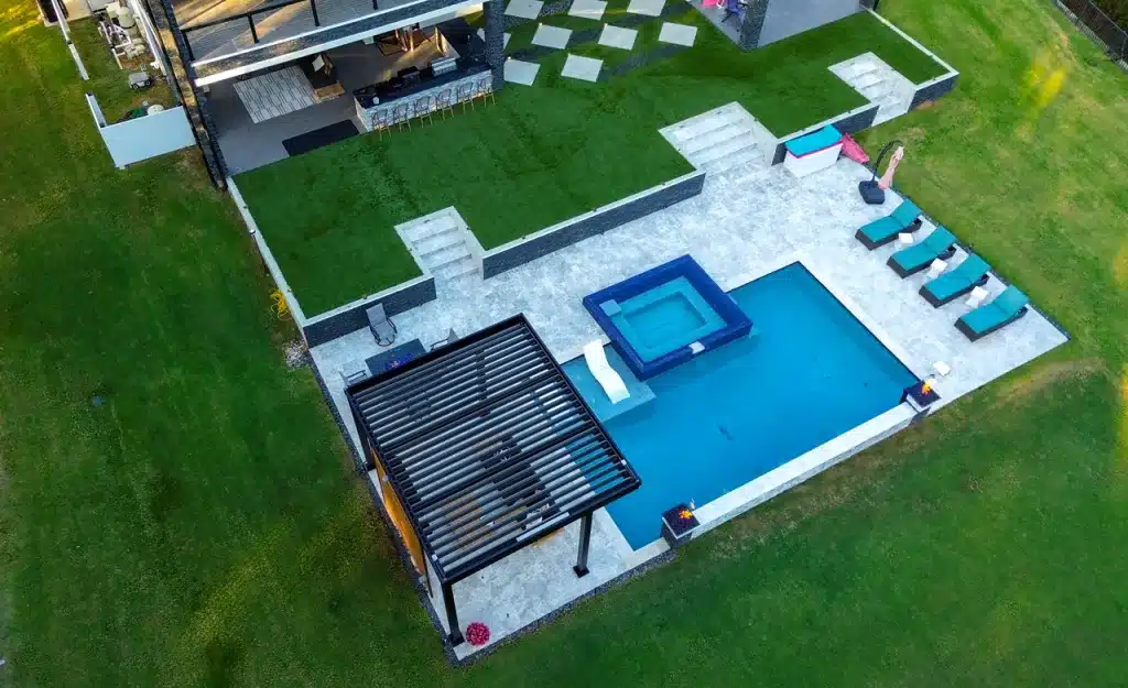 Aerial view (drone) of a black R-Blade pergola shading a luxury geometric pool and stone spa area with lounge chairs.