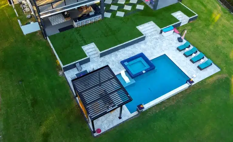 Aerial view (drone) of a black R-Blade pergola shading a luxury geometric pool and stone spa area with lounge chairs.