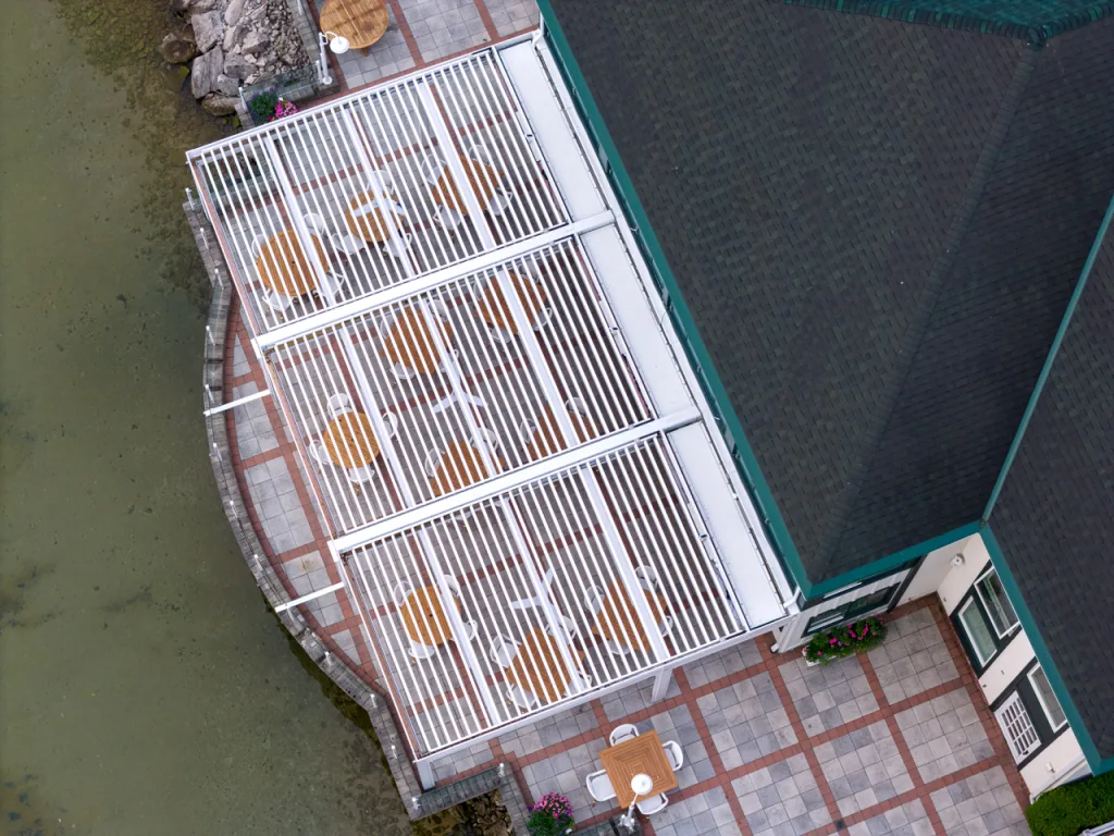View of a stylish outdoor dining area under a retractable louvered pergola, featuring white furniture, wood tables, and a retractable screen for privacy or weather protection.