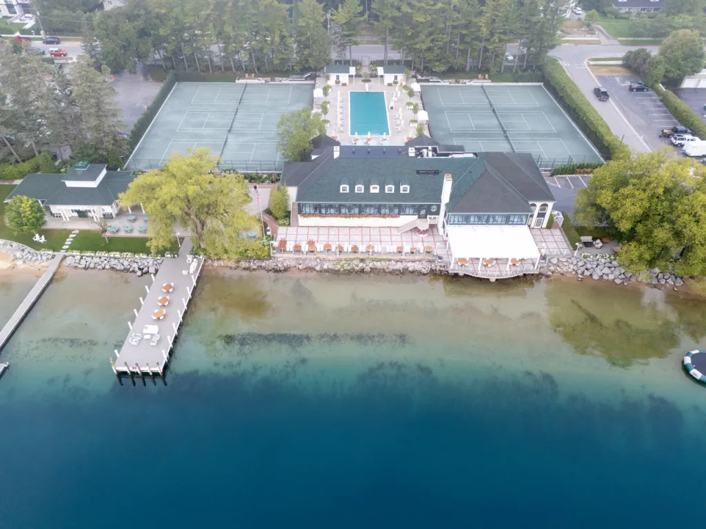 Stunning aerial view of a luxurious lakefront club featuring a restaurant, tennis courts, pool area, and a large dining deck with a white modern pergola in northern Michigan.