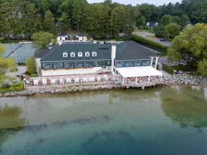Aerial view of a lakeside restaurant with an extensive outdoor dining patio under a white louvered pergola, featuring white wicker chairs and wood tables right on the water's edge.