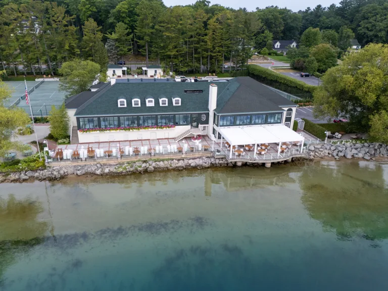 Aerial view of a lakeside restaurant with an extensive outdoor dining patio under a white louvered pergola, featuring white wicker chairs and wood tables right on the water's edge.