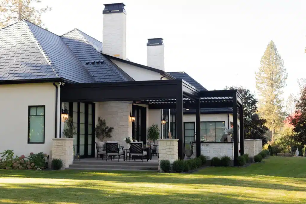 Luxury home exterior with Azenco R-Blade™ louvered pergola featuring a black aluminum frame and white stone columns over a paved patio.